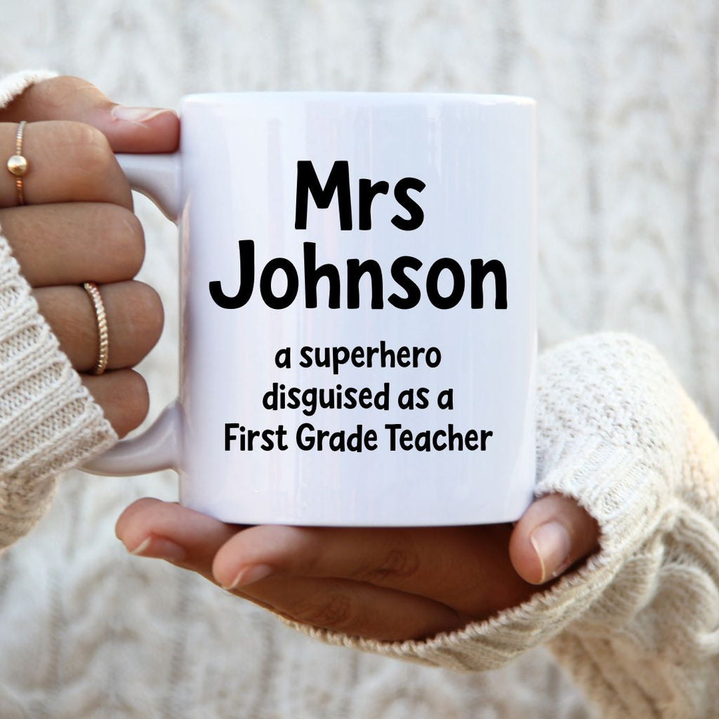 White mug with text 'Mrs Johnson, a superhero disguised as a First Grade Teacher' held by a person.