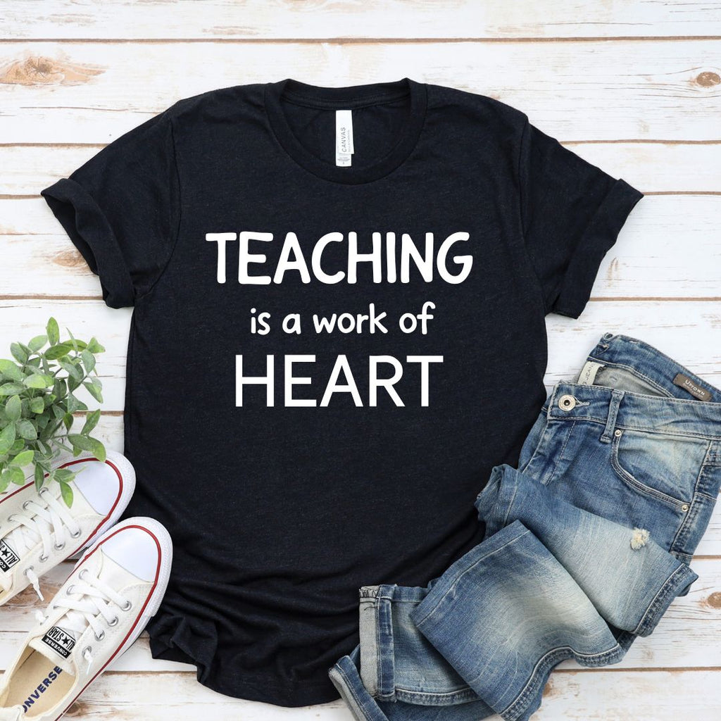 Black t-shirt with 'Teaching is a work of heart' text on a wooden surface with jeans and sneakers.