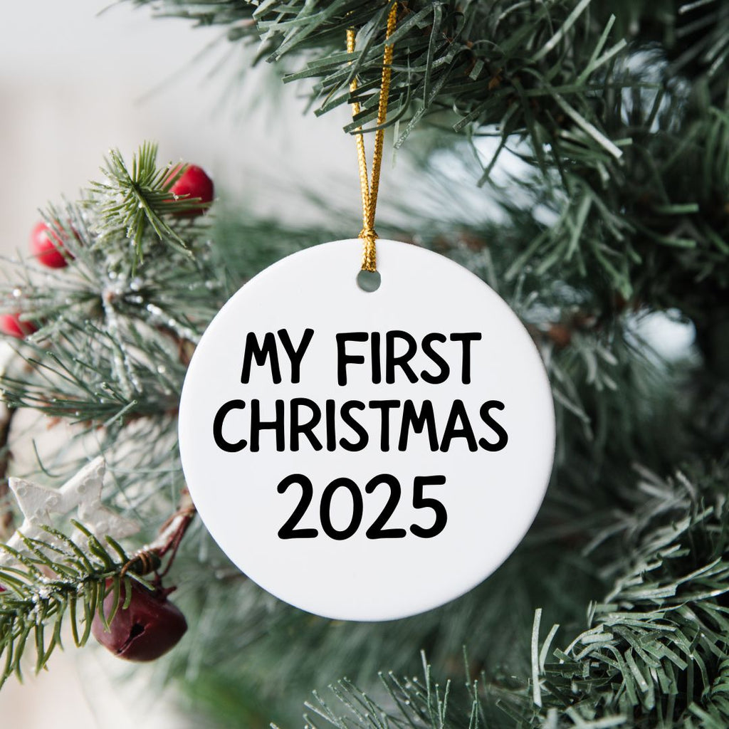 Round white ornament with 'My First Christmas 2025' text hanging on a Christmas tree.